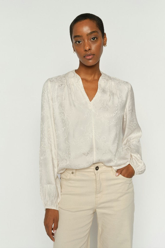 A women wearing An off-white long-sleeve blouse with delicate floral embroidery with cream jeans on a plain white background.