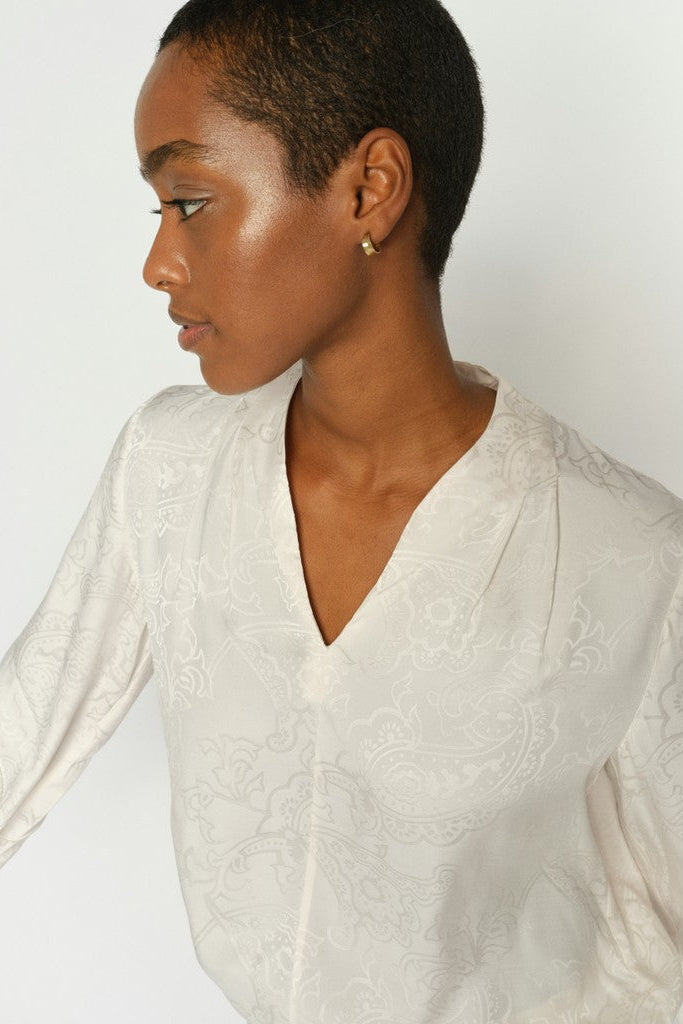 Close up side profile of woman wearing an off-white long-sleeve blouse with delicate floral embroidery, shown from the back against a plain white background.