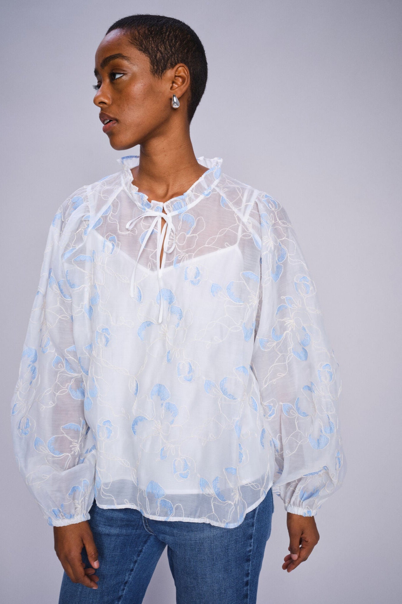 Model wearing white flowy blouse with voluminous sleeve  with blue abstract patterns on a white background paired with denim jeans. 