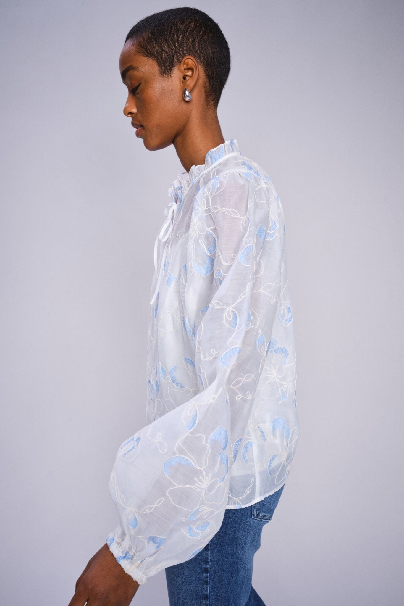 Model wearing white flowy blouse with voluminous sleeve  with blue abstract patterns on a white background paired with denim jeans. 