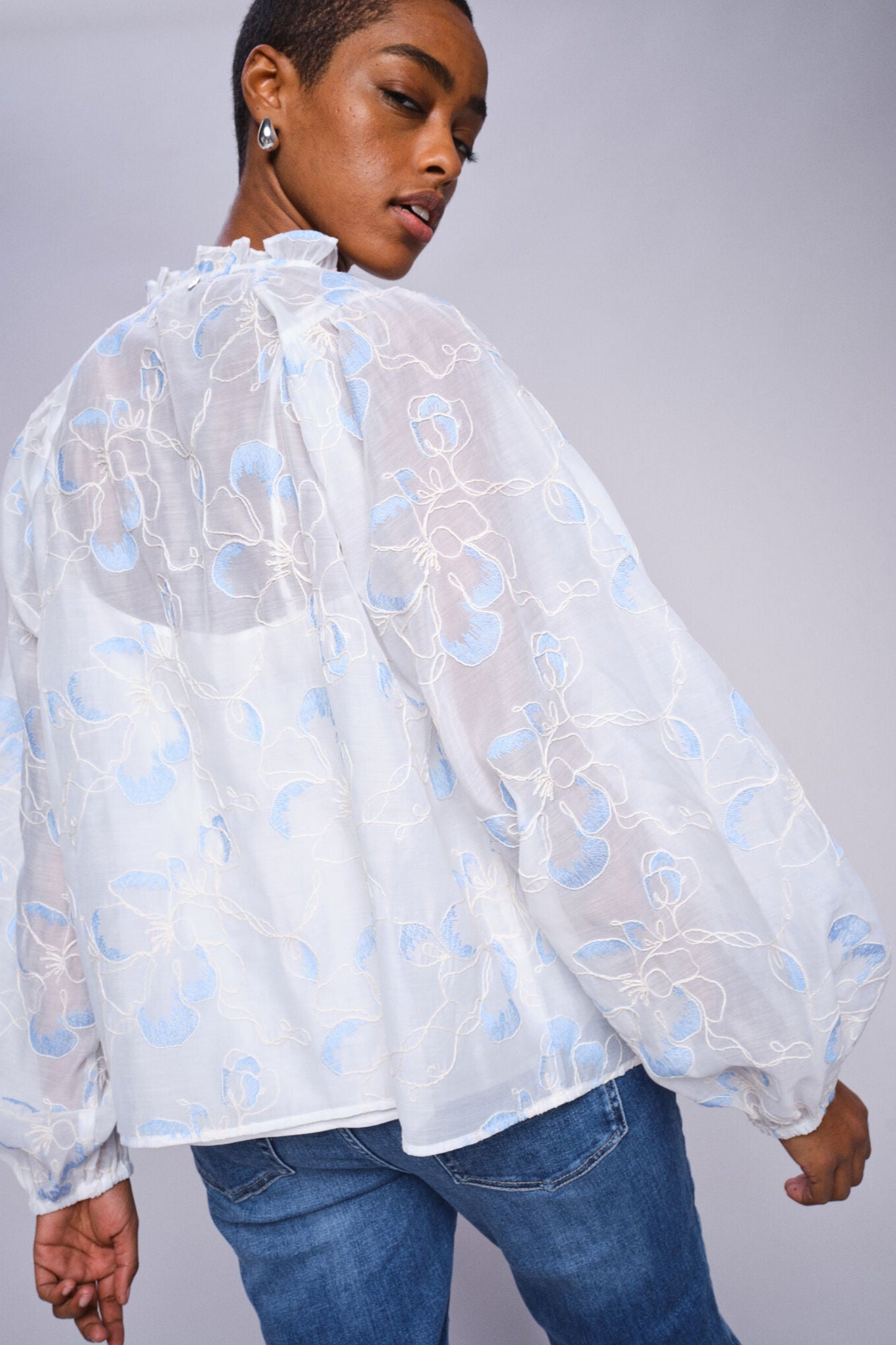 Model wearing white flowy blouse with voluminous sleeve  with blue abstract patterns on a white background paired with denim jeans. 