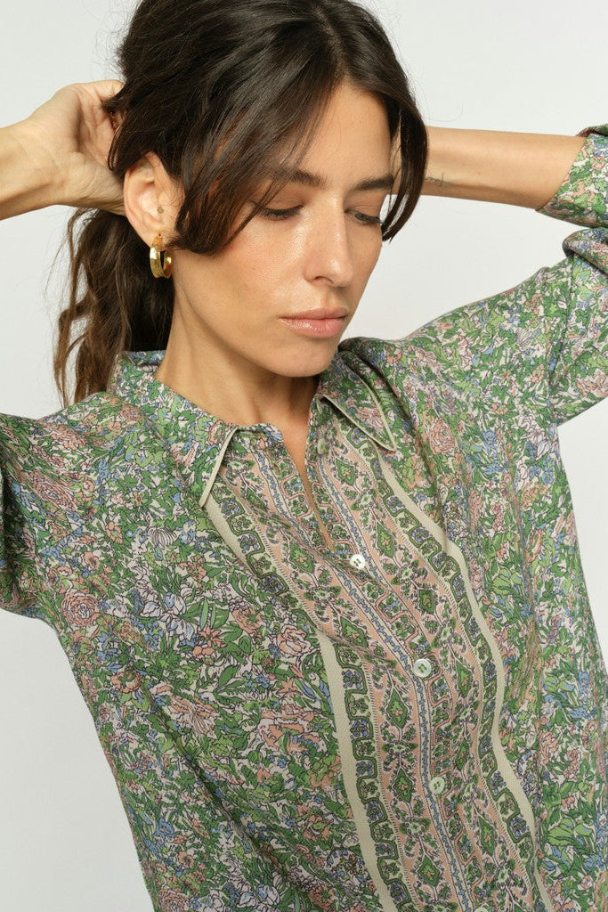 A woman wearing a green long-sleeve button-up shirt with a dense multicolor floral and paisley print, posing with her hands behind her head against a light gray background.