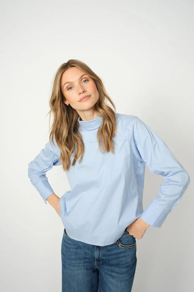 Woman wearing a light blue shirt with stand up collar and jeans on a white background