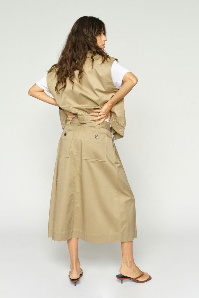 Woman wearing a beige waist coat  over a white tee shirt  with a matching beige midi skirt on a plain background