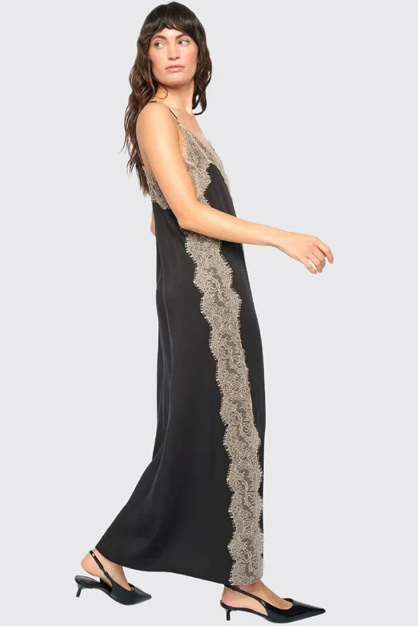 A woman modeling a sleeveless black maxi dress with textured beige lace panels down the sides, paired with black pointed-toe mule heels, against a solid light gray background.