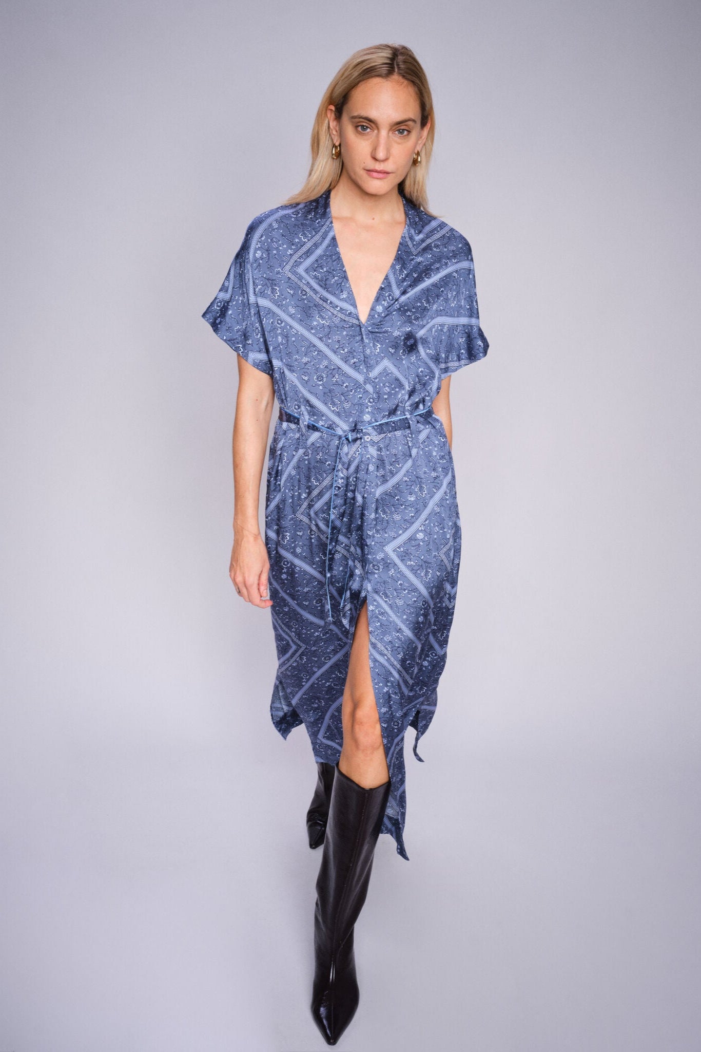 Model wearing a blue patterned dress with a slit on a gray background paired with black boots 