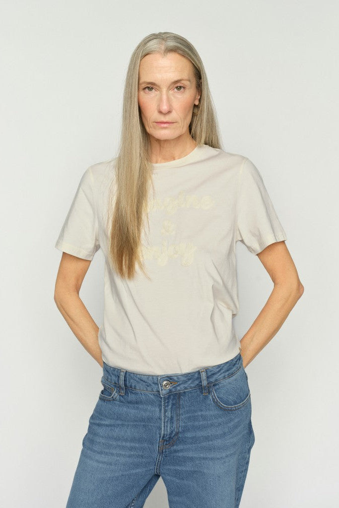 A woman with long blonde hair wearing a light beige short-sleeve t-shirt with tonal embroidered text, paired with blue jeans, against a plain light gray background.