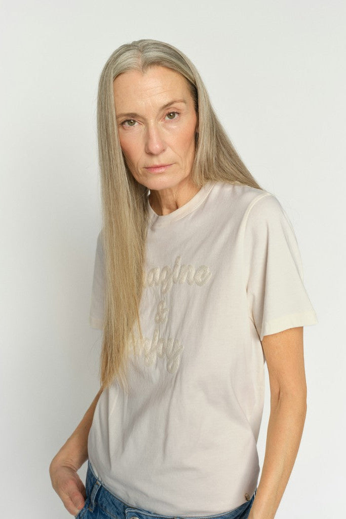 A woman with long blonde hair wearing a light beige short-sleeve t-shirt with tonal embroidered text, paired with blue jeans, against a plain light gray background.