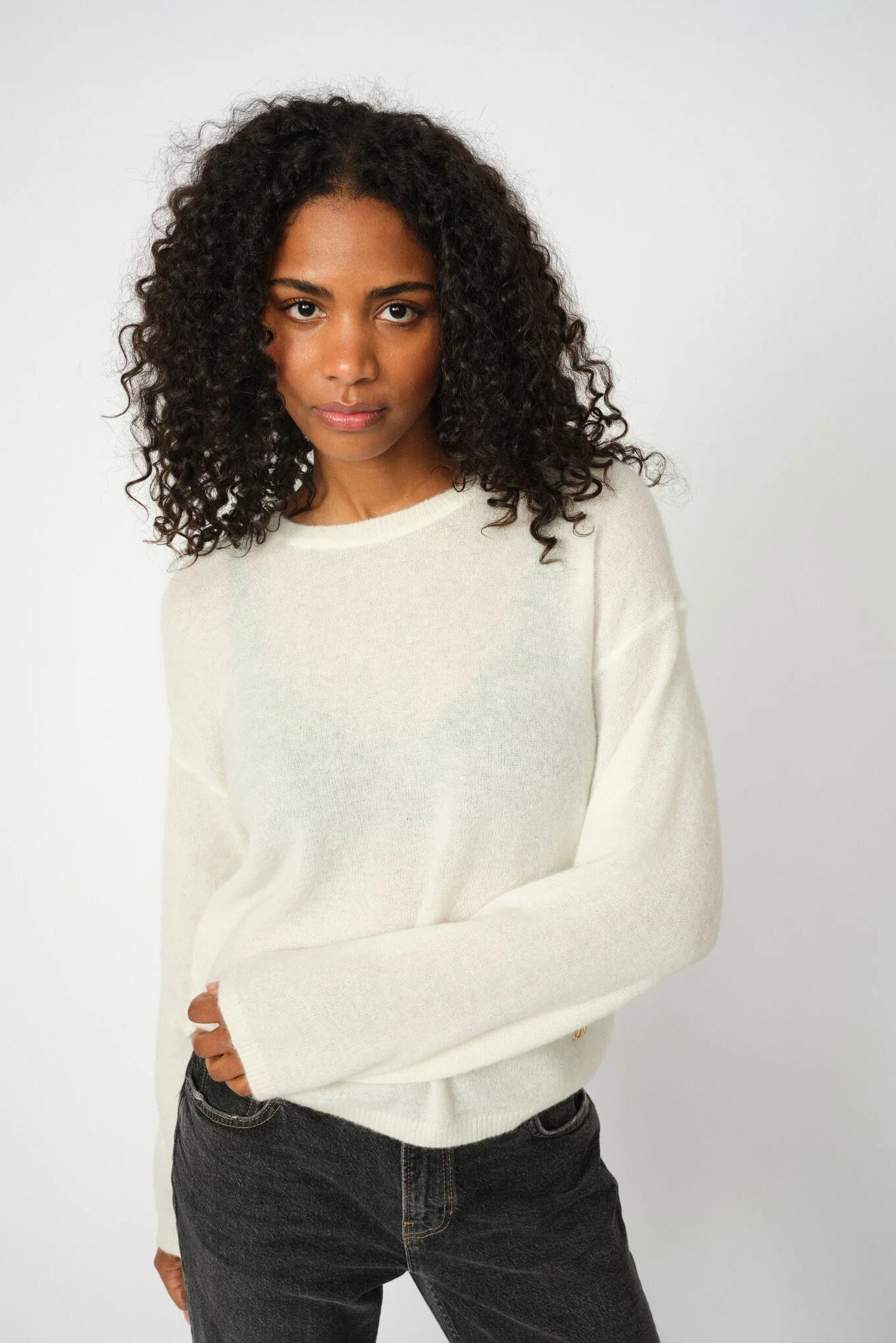 Woman wearing a white sweater and dark jeans against a plain background