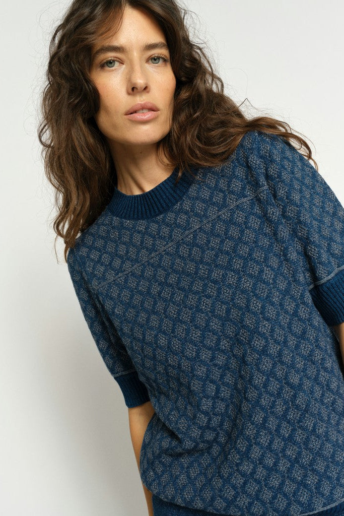 Close up of model showing details of a navy blue knit sweater with a textured geometric diamond pattern, featuring a crew neck, short sleeves with ribbed cuffs, and a ribbed hem, on a white background.