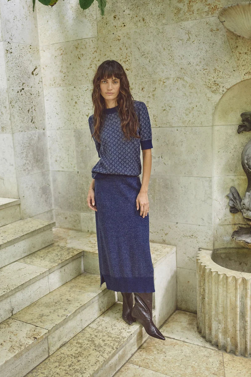 Woman in a blue short sleeve sweater with geometric print and blue skirt standing on stone steps with a fountain in the background