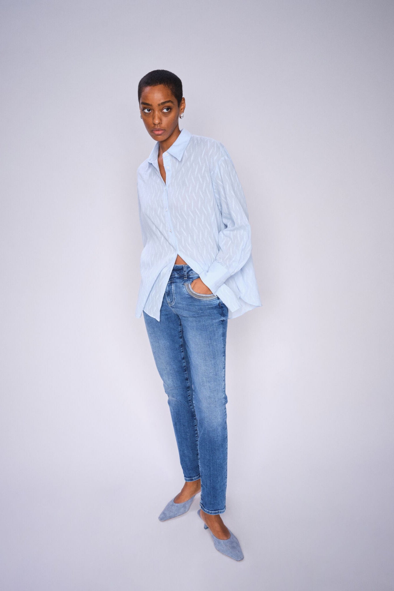 Person wearing a light blue shirt and  mid blue slim leg jeans against a plain background
