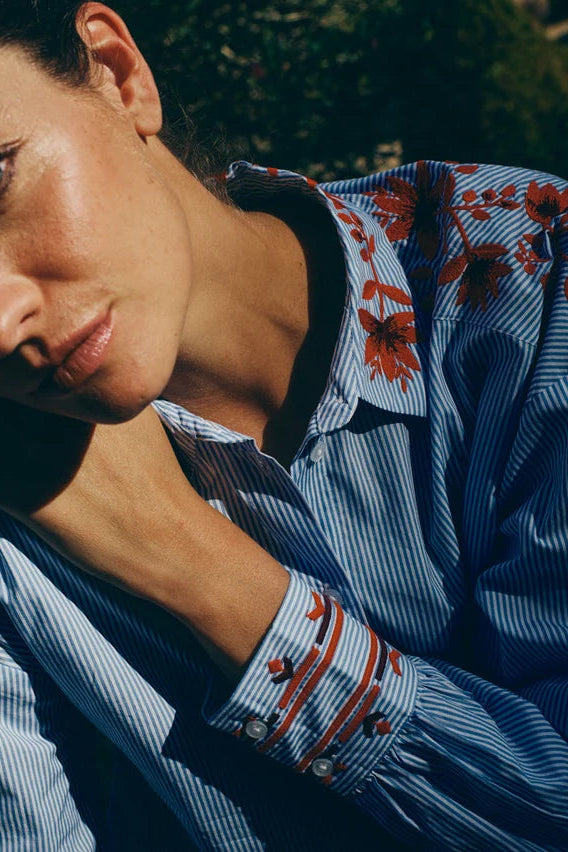 Woman wearing a blue shirt with red floral embroidery sitting outdoors.