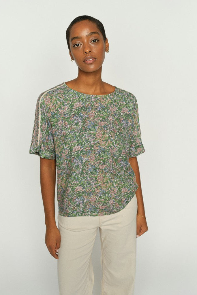Model wearing a floral print short sleeve shirt in green, pink, and beige tones, featuring contrasting light beige piping along the shoulder seams and neckline.  She is wearing cream pants and is shown on a plain background. 