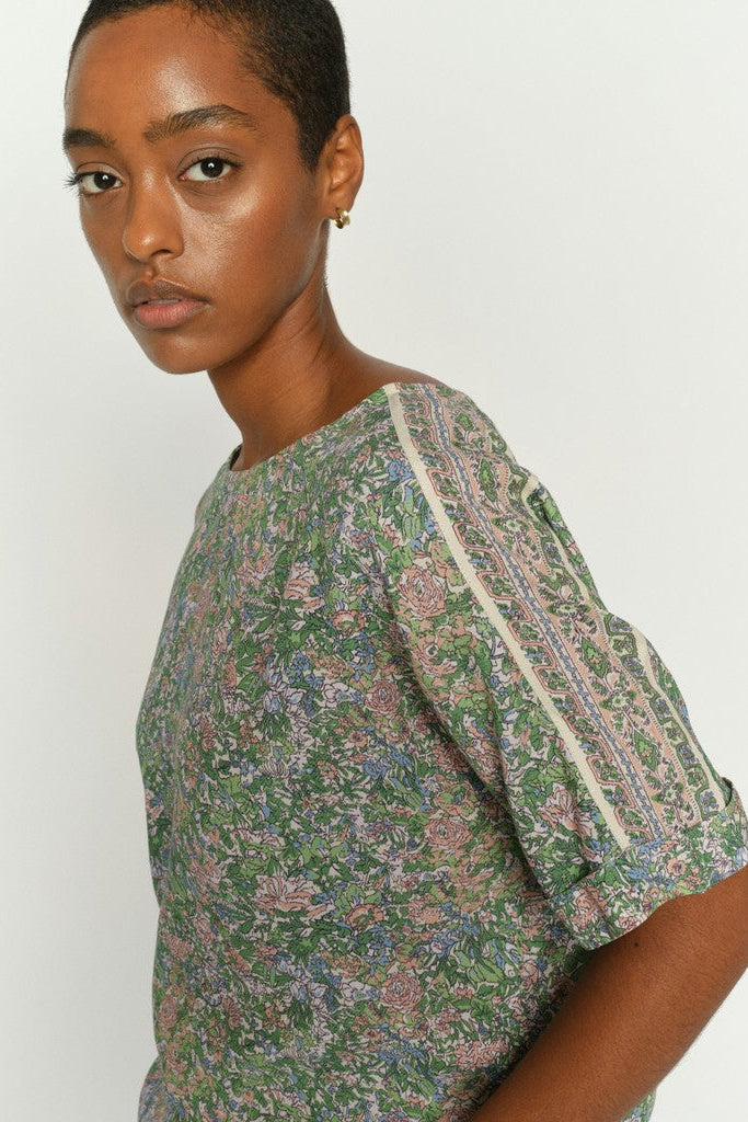 Model wearing a floral print short sleeve shirt in green, pink, and beige tones, featuring contrasting light beige piping along the shoulder seams and neckline. Shown on a plain background. 