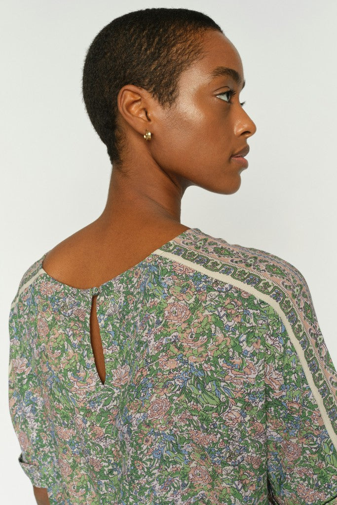 Women shown from back modelling a short-sleeve top with an all-over dense floral print in green, pink, and beige tones, featuring contrasting light beige piping along the shoulder seams and neckline. Shown on a plain background. 