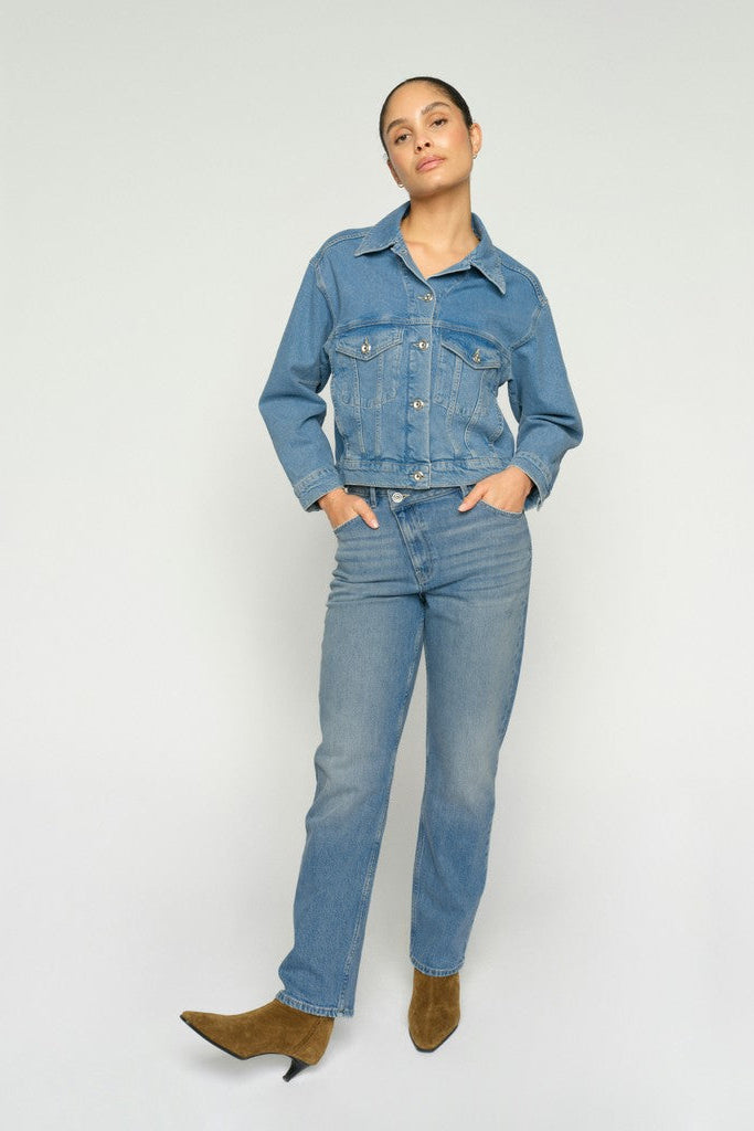 A woman modeling a light blue denim jacket and matching blue jeans against a plain light gray studio background.