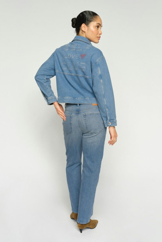 A woman shown from the back modeling a light blue denim jacket and matching blue jeans against a plain light gray studio background.