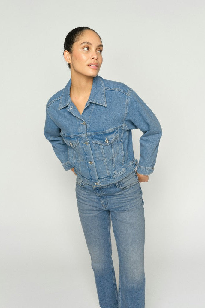 A woman modeling a light blue denim jacket and matching blue jeans against a plain light gray studio background.