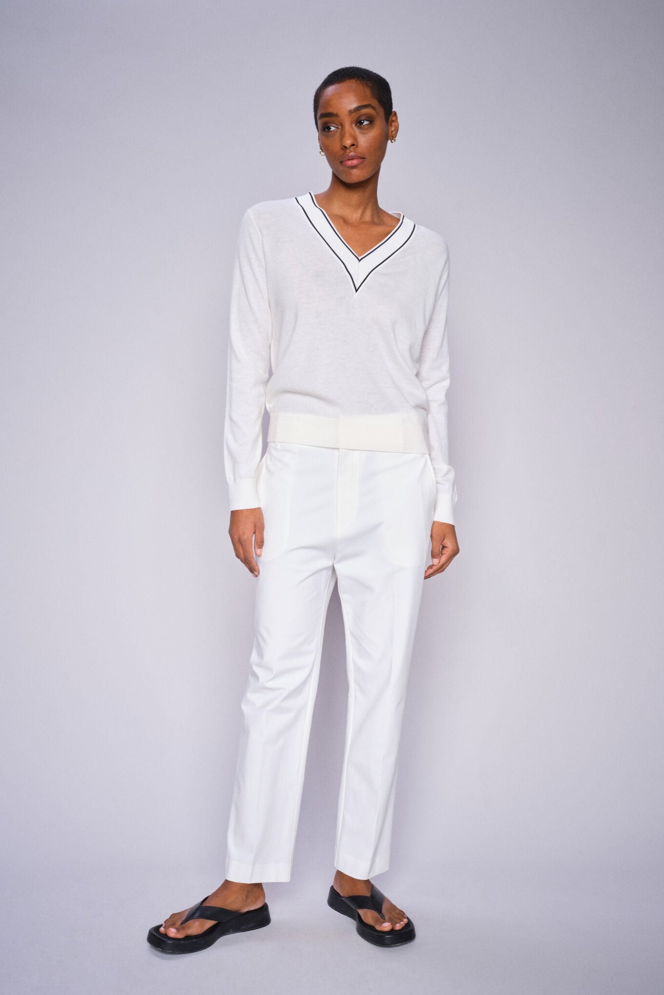 Woman wearing ecru v neck sweater and ecru pants with black sandals on a grey background. 