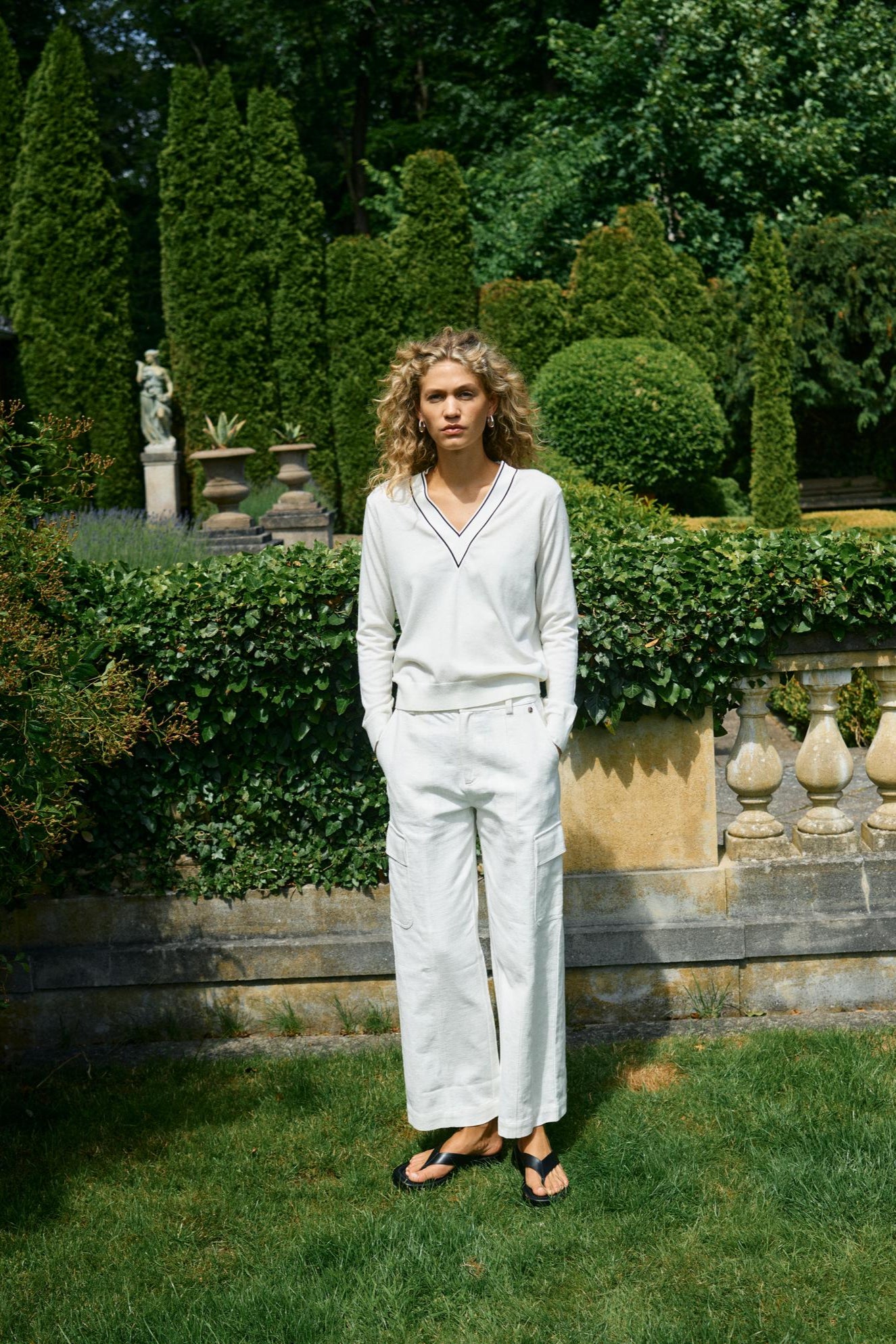 Woman wearing ecru v neck sweater and ecru pants with black sandals  in a garden with manicured hedges and statues.