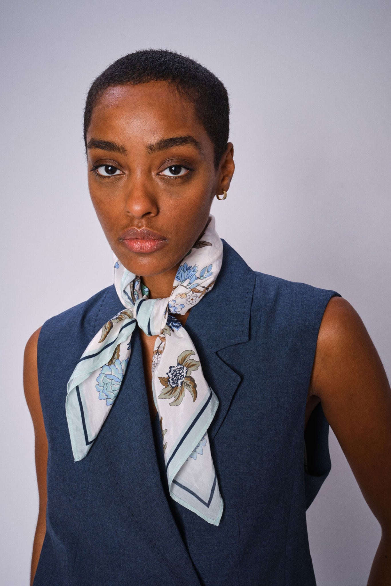 Model wearing Floral square scarf with ecru base and blue brown and green floral print all over tied around her neck with blue top. 