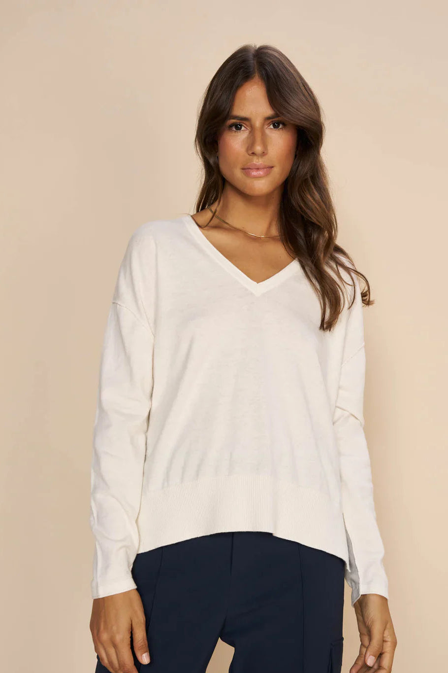 Woman wearing a white v neck long sleeve sweater and dark pants against a beige background
