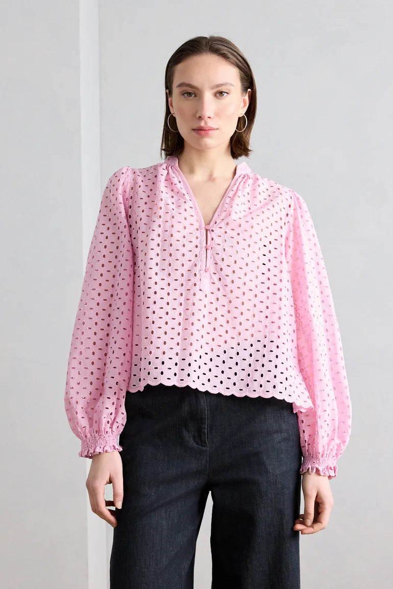 Woman wearing a pink broderie blouse with long sleeves and wide leg pants against a plain background