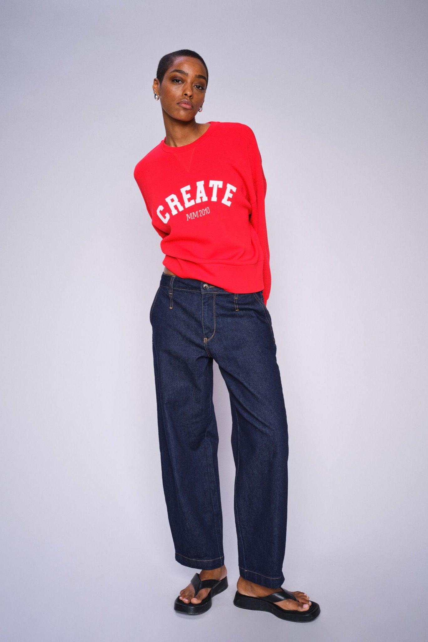 Person wearing a red sweatshirt with 'CREATE' text and blue chino style jeans on a gray background