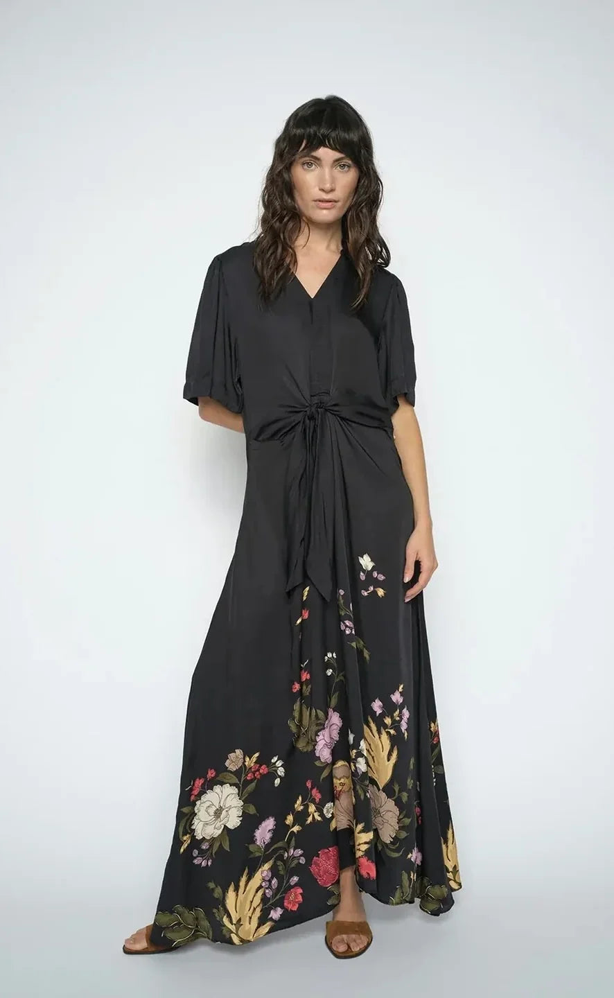 A women wearing a  black wrap-style maxi dress with short flutter sleeves, a V-neckline, a front waist tie, and a floral print concentrated along the lower skirt, shown on a plain white background.