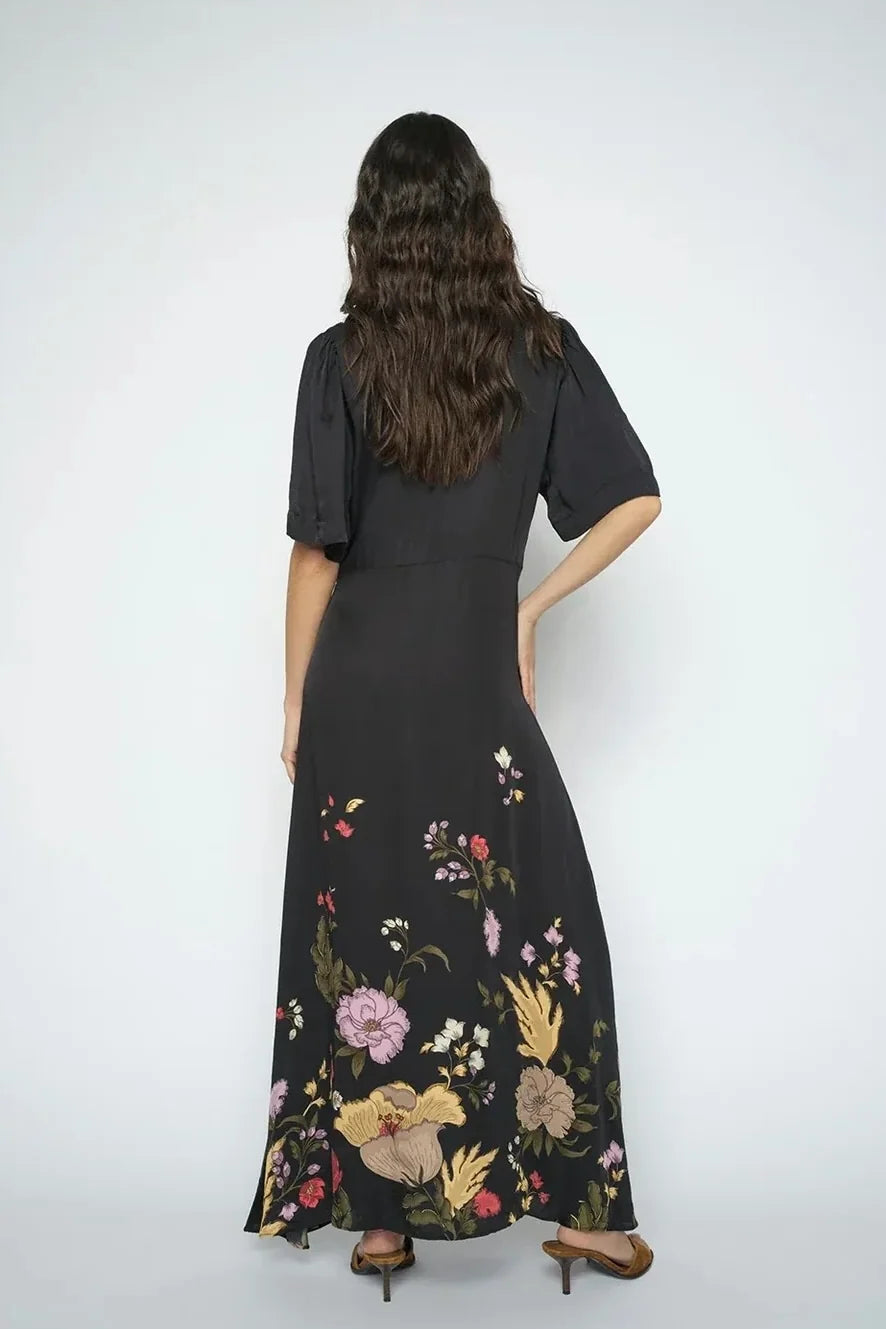 A women shown from the back wearing a  black wrap-style maxi dress with short flutter sleeves, a V-neckline, a front waist tie, and a floral print concentrated along the lower skirt, shown on a plain white background.