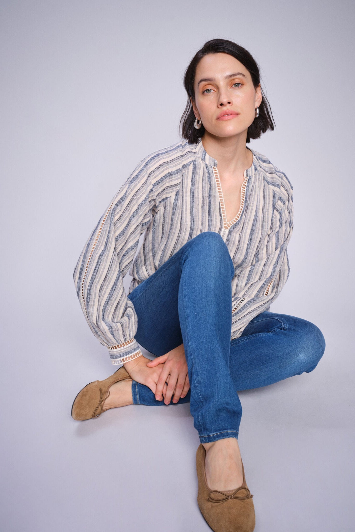 Woman wearing a blue and white stripe cotton linen shirt and blue jeans sitting on a white background