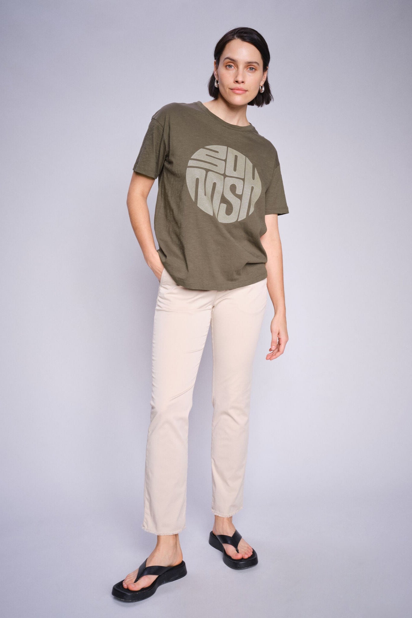 Woman wearing a olive green t-shirt with a logo and white pants on a gray background
