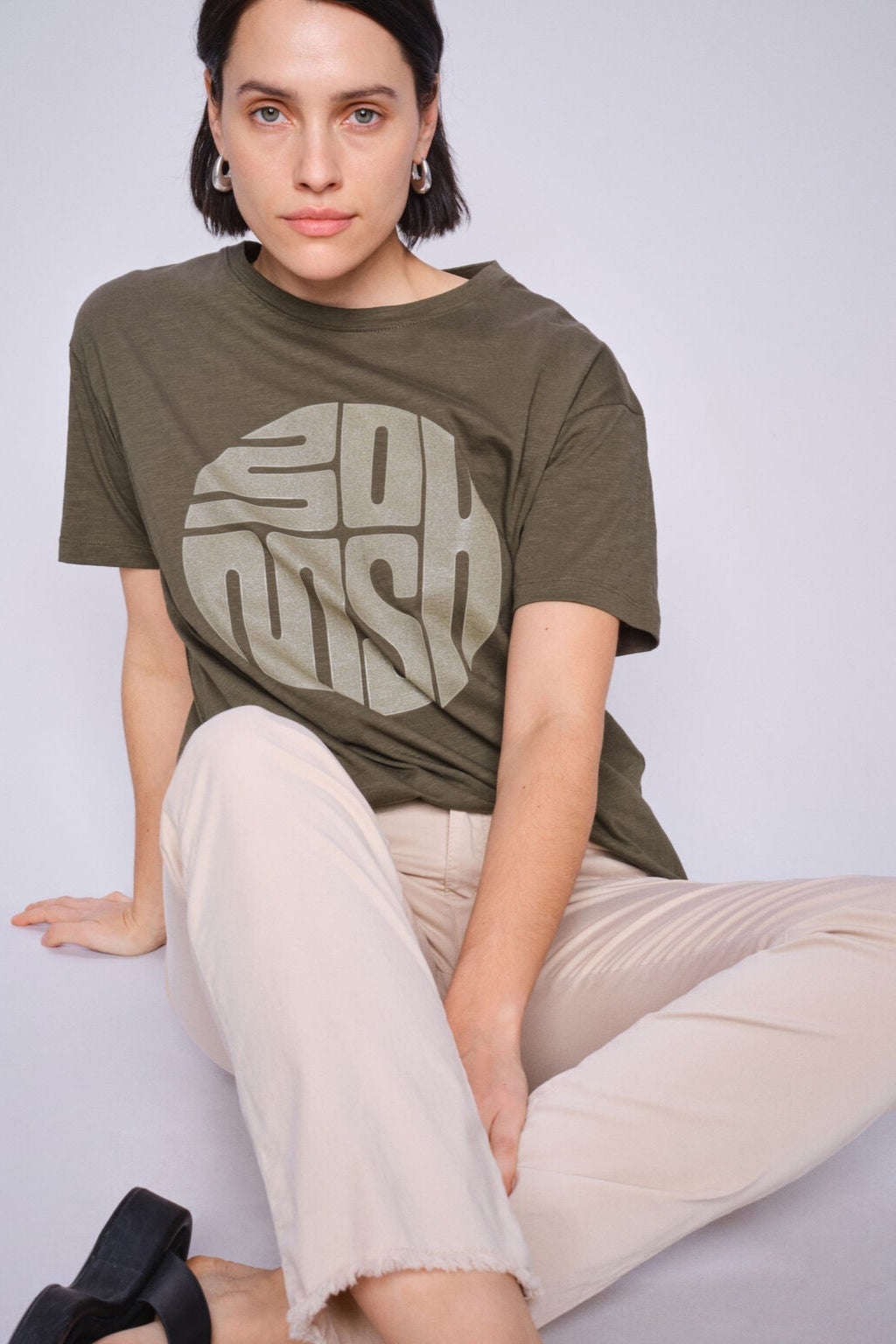 Model wearing an olive green t-shirt with a logo, sitting against a plain background