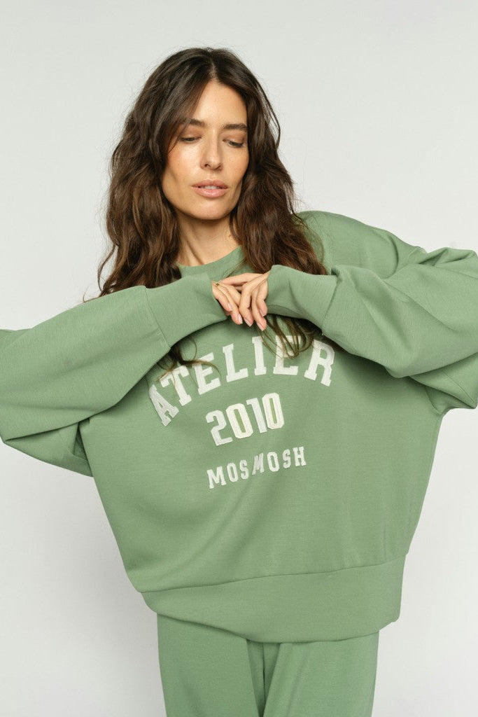 A women with long brown hair modelling a sage green crewneck sweatshirt with 'ATELIER 2010 MOS MOSH' printed in white on the front and matching sage green track pants on a clean white background.
