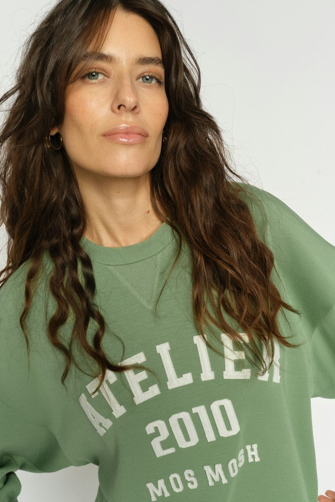 A women with long brown hair modelling a sage green crewneck sweatshirt with 'ATELIER 2010 MOS MOSH' printed in white on the front, displayed on a clean white background.