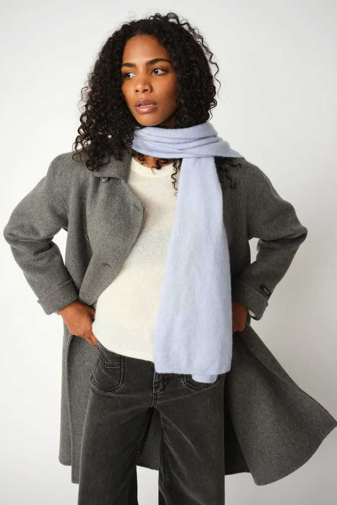 A woman modeling a light blue scarf wrapped around her neck, paired with a gray coat, cream sweater, and dark gray corduroy pants against a plain light gray background.
