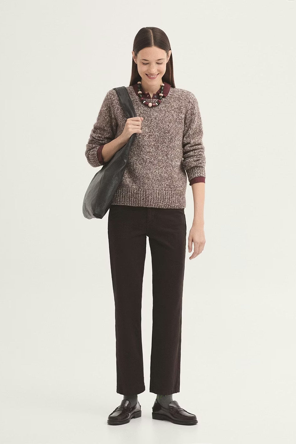 Person wearing dark brown pants and a red sweater on a white background with black loafers and a black handbag. 