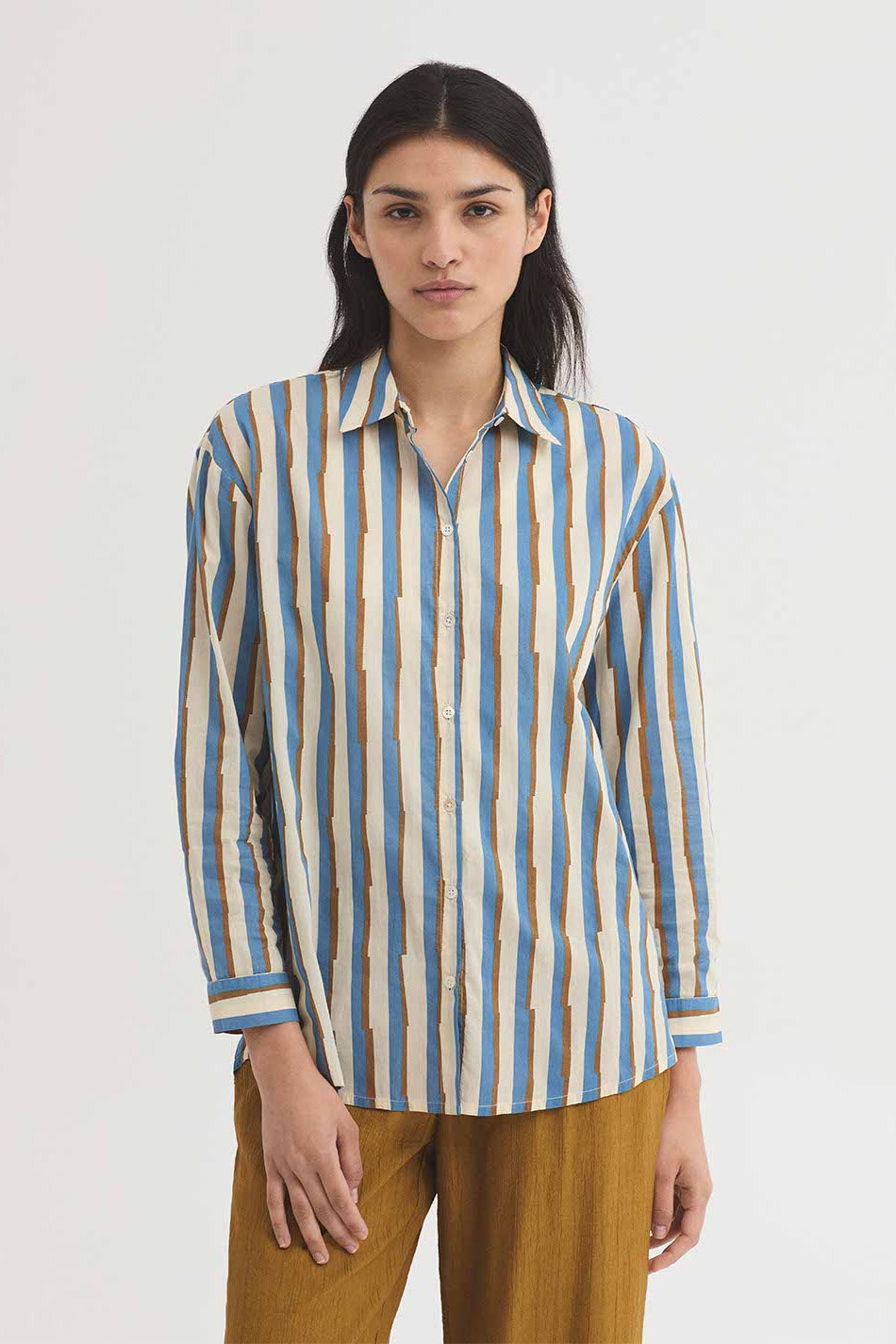 A woman modeling a long-sleeve button-up shirt with vertical stripes in blue, beige, and brown, paired with brown trousers, against a plain light gray background.