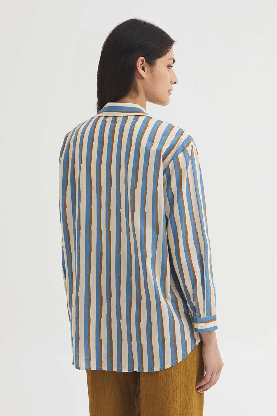 A woman shown from the back wearing a long-sleeve button-up shirt with vertical blue, beige, and white stripes, paired with brown trousers, against a plain white studio background.