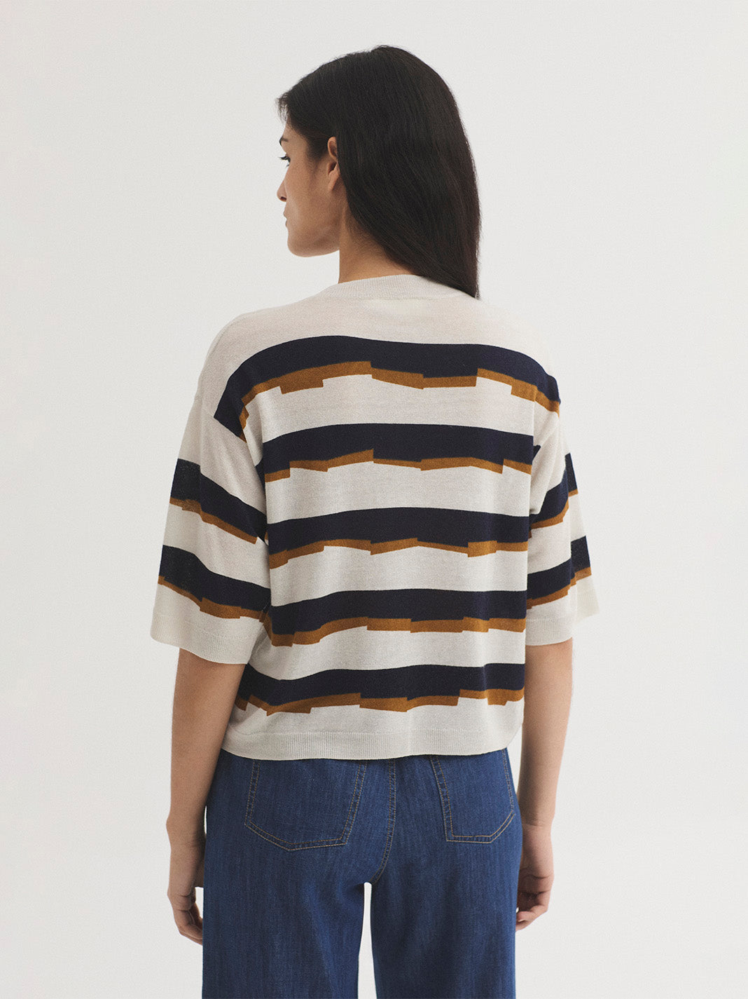 A woman viewed from the back wearing a short-sleeve knit top with horizontal navy, brown, and off-white stripes, paired with blue jeans, against a plain light background.