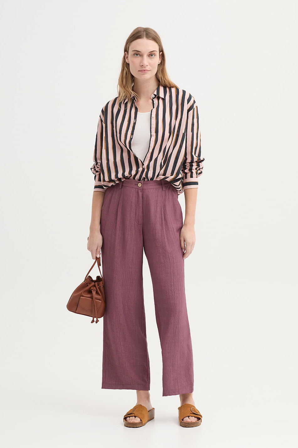 A woman stands against a plain light background wearing a beige and black vertically striped button-up shirt, mauve wide-leg trousers, tan sandals, and holding a small brown handbag.