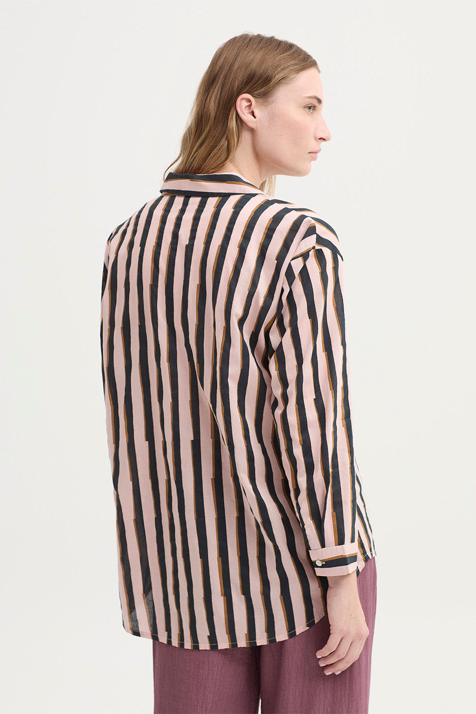 A woman shown from the back wearing a long-sleeve button-up shirt with vertical black stripes on a warm beige background, paired with mauve-purple trousers, against a plain light gray studio background.