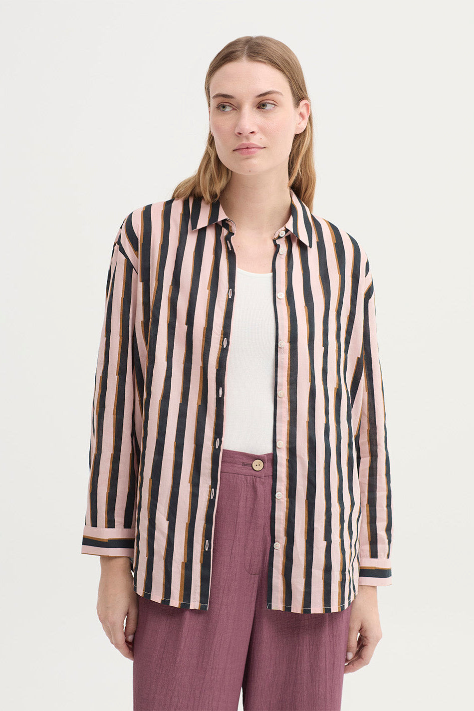 A woman modeling a long-sleeve button-up shirt with vertical stripes in black, beige, and dusty pink, worn open over a white top and mauve trousers, against a plain light background.