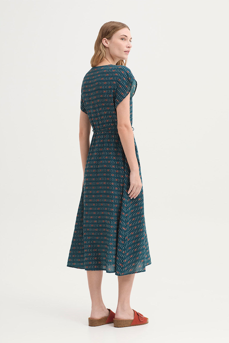 A woman shown from the back wearing a dark teal wrap-style dress with a wavy abstract pattern in white and rust orange, photographed against a plain light background.