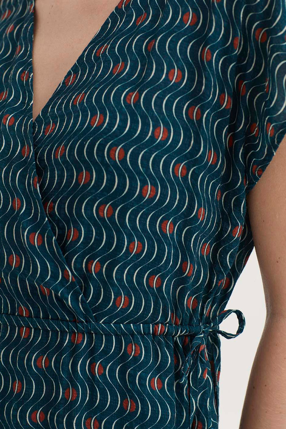 Close-up of a wrap-style dress with a dark teal blue base, featuring a repeating pattern of white wavy lines and small red crescent motifs, worn by a model.