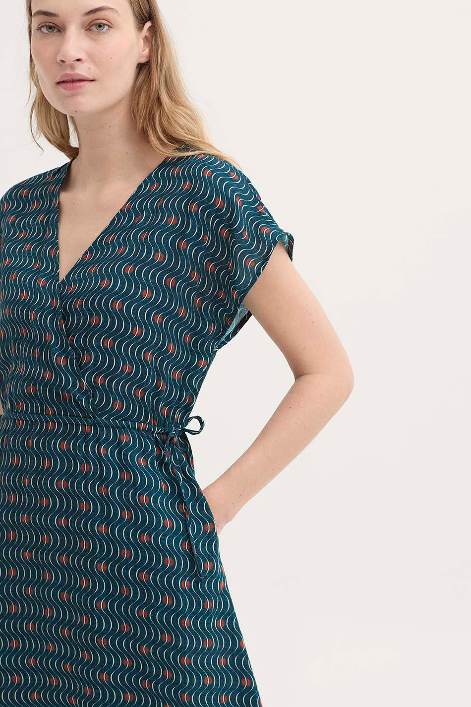 A woman wearing a dark teal wrap-style dress with a wavy abstract pattern in white and rust orange, photographed against a plain light background.