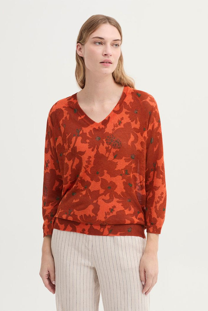 A woman modeling a rust-orange long-sleeve V-neck top with a dark floral camouflage pattern, paired with light beige pinstriped trousers, against a plain off-white studio background.