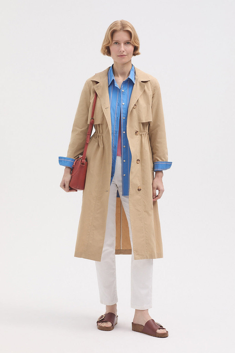 Model wearing a blue striped shirt over a pink top with white trousers and beige trench coat and sandals. 