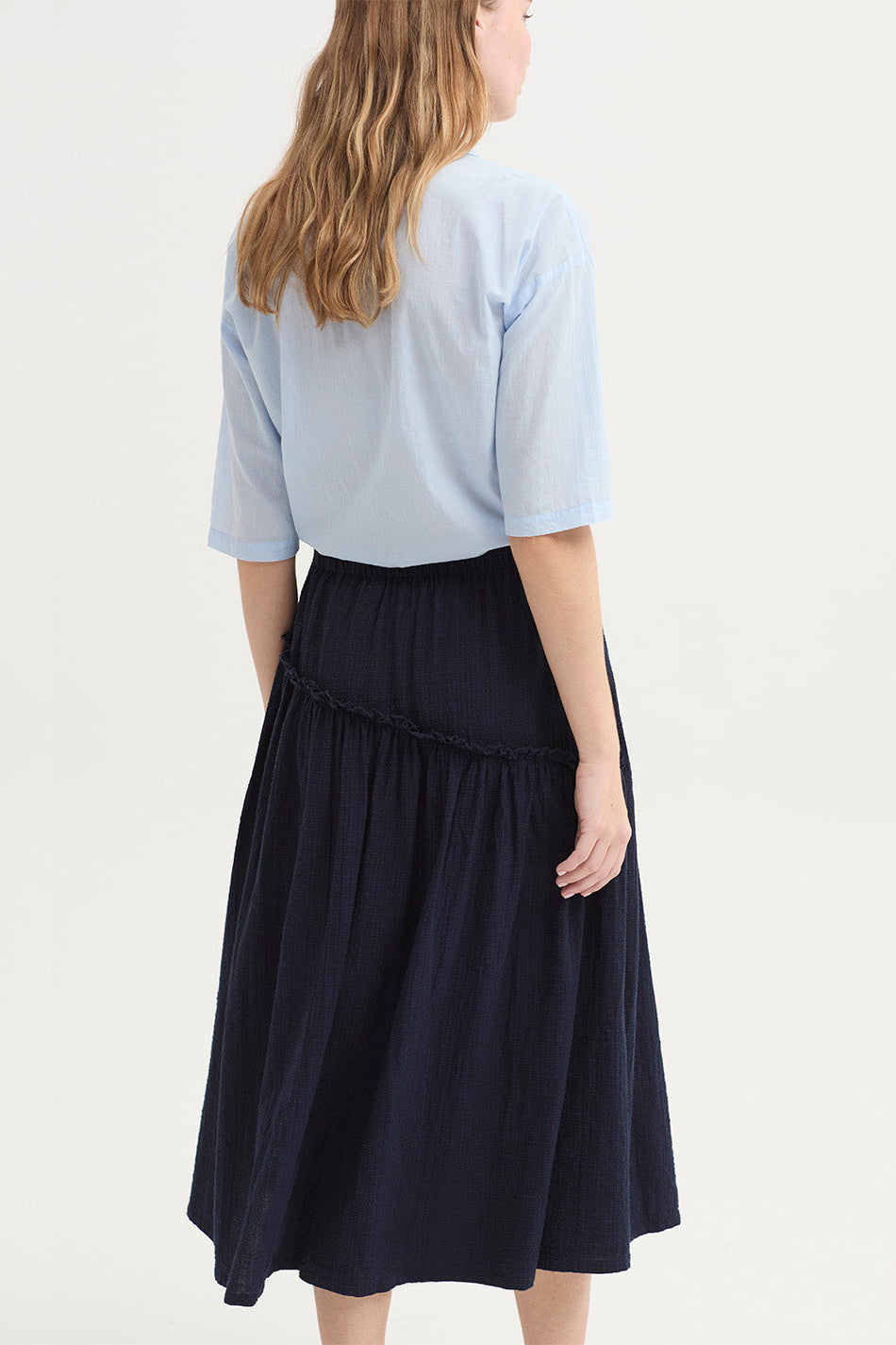 A woman shown from the back wearing a light blue short-sleeve blouse and a dark navy tiered midi skirt against a plain light background.
